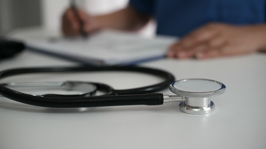 A stethoscope on a desk while a health worker writes notes in the background
