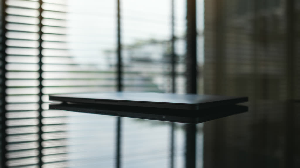 A closed laptop on a dark table beside window blinds