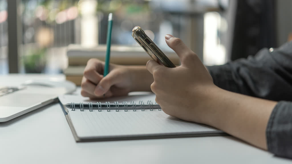 A person holding a phone while writing notes in a notebook
