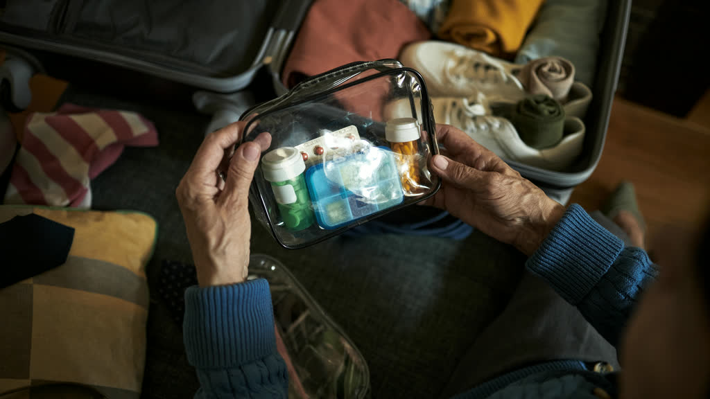 Hands holding a clear toiletry bag with pill bottles and a weekly pill organiser over an open suitcase