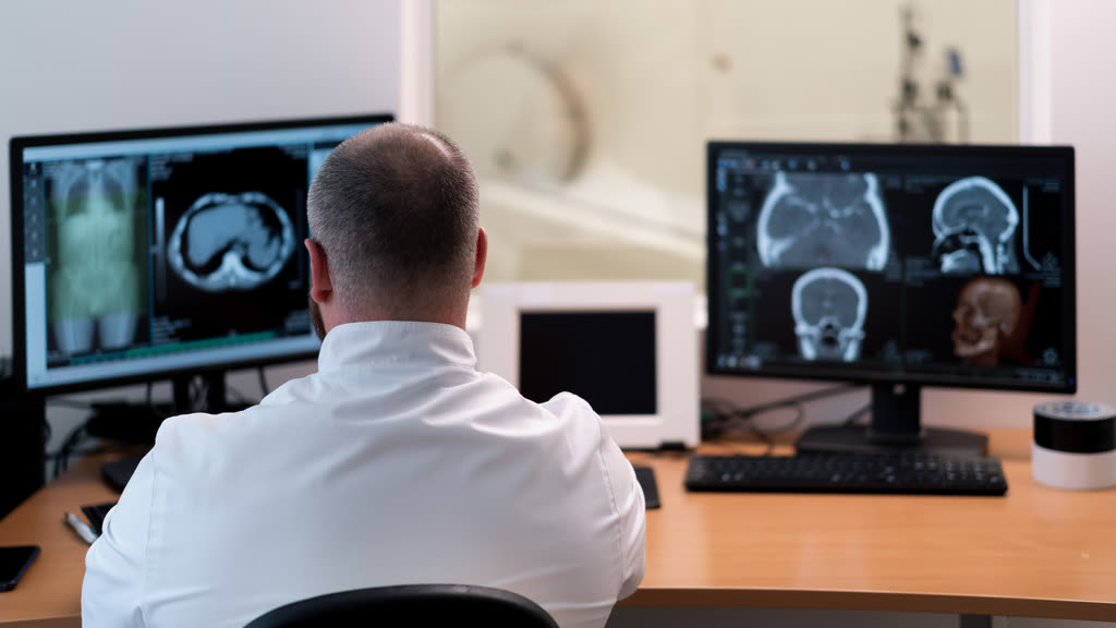A doctor, seen from behind, reviewing brain scans on two monitors