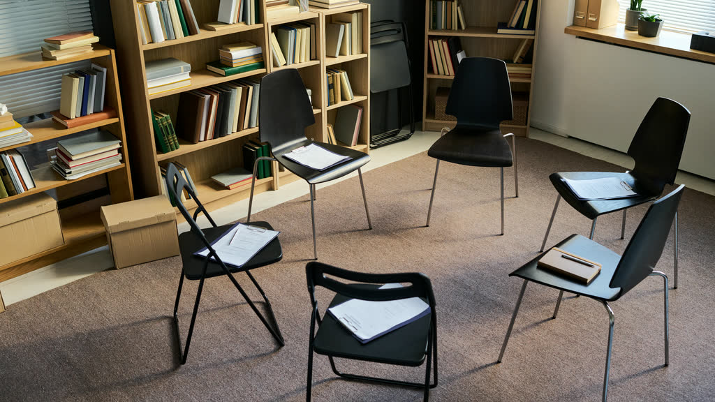 A circle of empty chairs in a room with bookshelves, set up for a group session